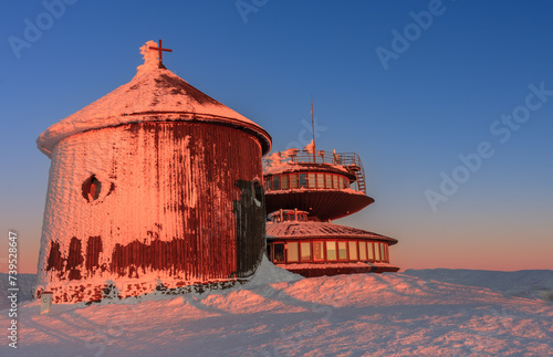 Winter, sunrise time, wooden Roman catholic chapel and disc shaped meteorological observatory in snezka, mountain on the border between Czech Republic and Poland.