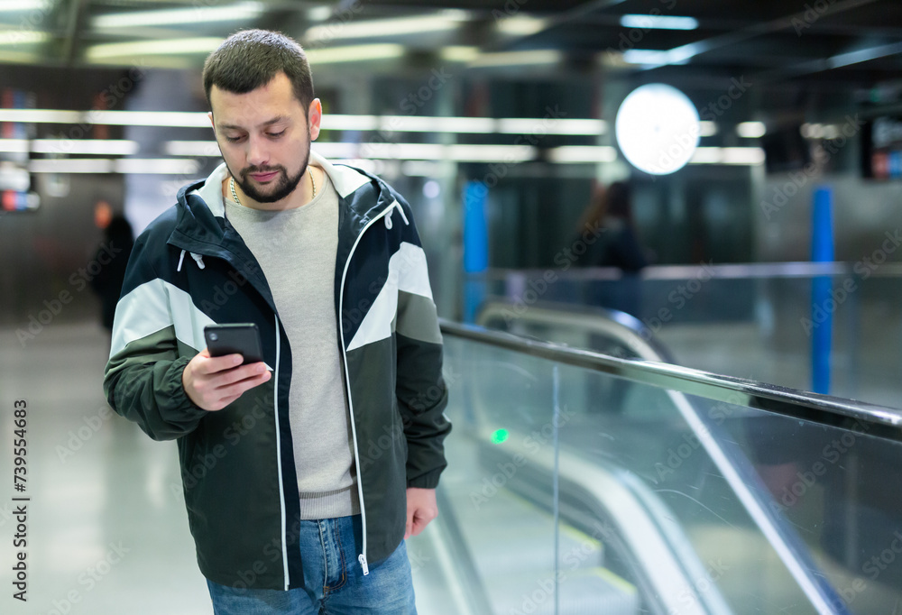 Portrait of positive bearded guy traveler standing on empty subway station with smartphone in hands, waiting for train ..