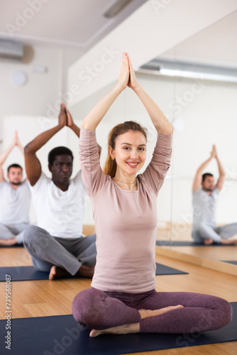 Positive young woman sitting in Padmasana position with her hands clasped in namaste gesture above head during group yoga class in fitness room