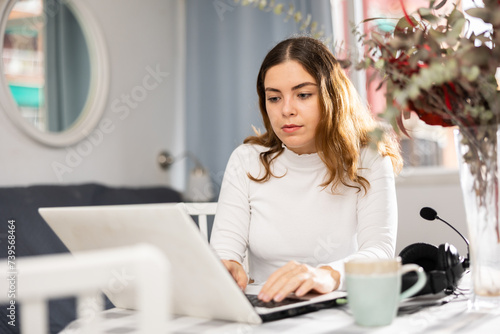 Young caucasian woman sitting at table at home in front of laptop and typing.