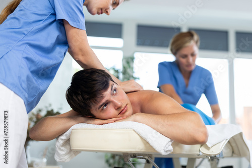 Massage in four hands. Two unrecognizable female masseuses perform stroking and rubbing of muscles of back and shoulders to young guy client of spa salon