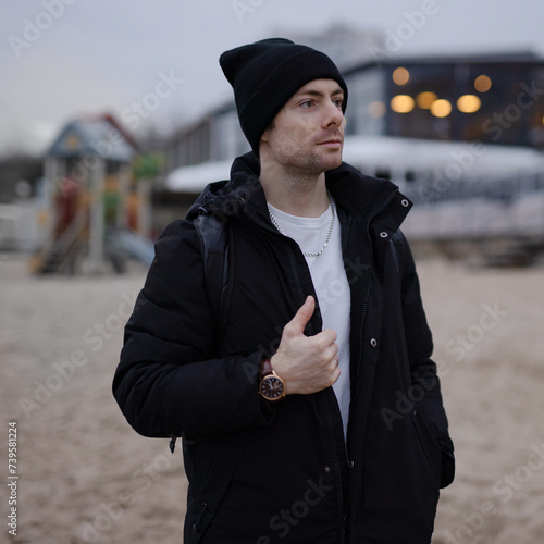 Young man in black winter jacket and hat on the beach