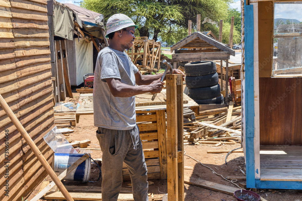african american worker making a wendy house, wooden shed outdoors, he is hammering a pallet to make planks african american worker making a wendy house, wooden shed outdoors, he is hammering a pallet to make planks