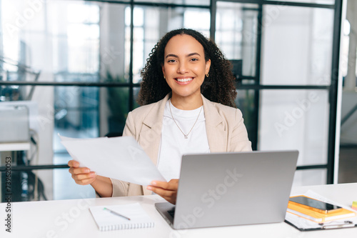 Attractive positive beautiful curly haired hispanic young woman in stylish elegant clothes, secretary, business lady, working with documents while sitting at her workplace in the modern office, smile