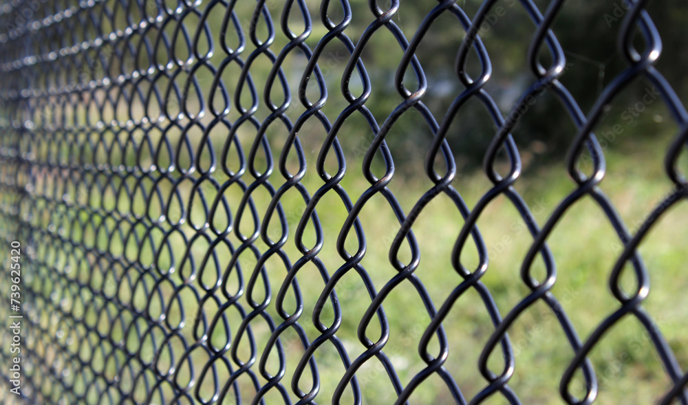 Fototapeta premium Close up view of a black chain-link fence with blurred greenery in the background
