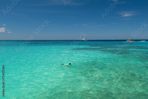 Man swimming in crystal clear shallow turquoise ocean water, deep blue sky and white yacht on the horizon. Saona Island, Dominican Republic. Wide angle shot.