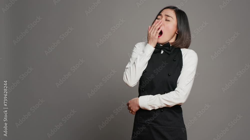 Exhausted waitress with apron yawning over grey background, feeling overworked and dealing with burnout after multiple restaurant events. Asian catering worker feeling tired in studio. Camera A. Exhausted waitress with apron yawning over grey background, feeling overworked and dealing with burnout after multiple restaurant events. Asian catering worker feeling tired in studio. Camera A.