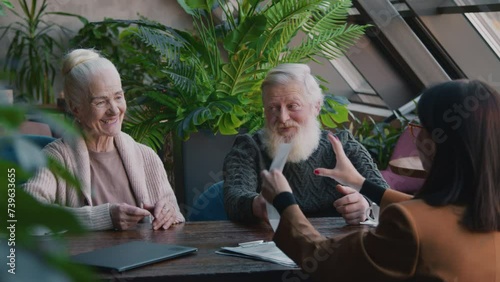 Medium shot of senior Caucasian man with grey beard talking to smiling wife while signing legal documents, discussing terms, then giving paperwork back to female broker and shaking hands photo