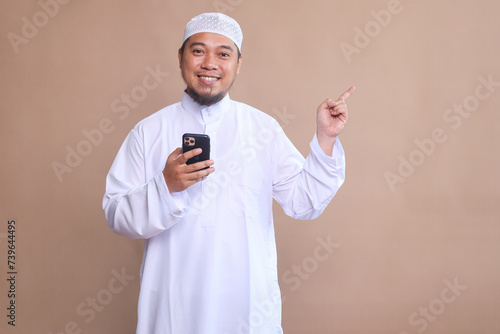 Smiling Asian Muslim man holding mobile phone and pointing away at copy space over beige background