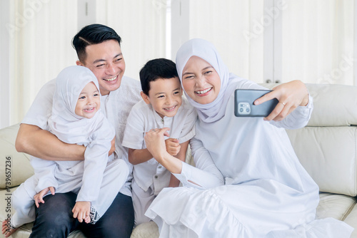 Happy muslim family making a video call or taking a selfie while sitting together on the couch during Eid Mubarak in the living room