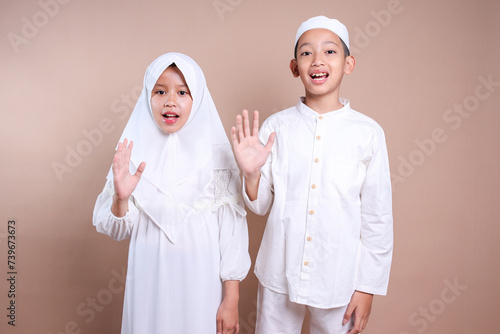 Two young Asian muslim little kids waving hands say hi or goodbye over beige background