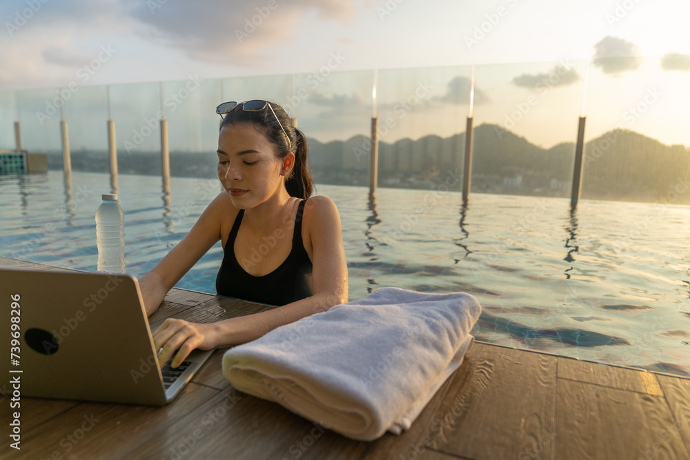 Beautiful white woman wearing a black bikini relaxing in swimming pool roof top at luxury hotel spa using laptop to check email, working anywhere.