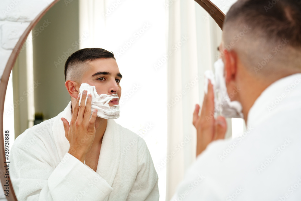 Reflection of a young man shaving in bathroom mirror close up
