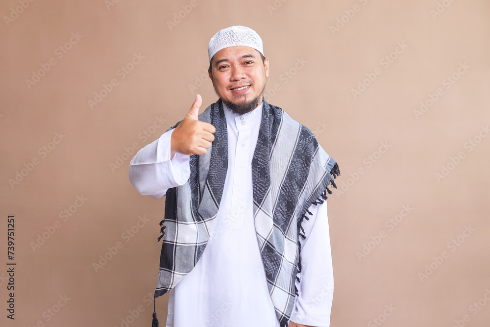 Asian muslim man feeling proud, carefree, confident and happy, smiling positively with thumbs up against beige background Asian muslim man feeling proud, carefree, confident and happy, smiling positively with thumbs up against beige background