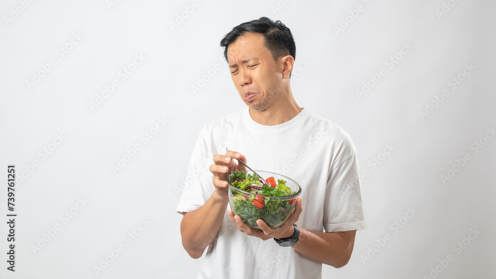 Portrait of an Indonesian Asian man, wearing a white T-shirt, joyfully posing while eating salad from a large transparent bowl, isolated against a white background. Portrait of an Indonesian Asian man, wearing a white T-shirt, joyfully posing while eating salad from a large transparent bowl, isolated against a white background.