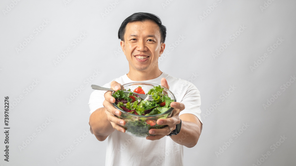 Portrait of an Indonesian Asian man, wearing a white T-shirt, joyfully posing while eating salad from a large transparent bowl, isolated against a white background. Portrait of an Indonesian Asian man, wearing a white T-shirt, joyfully posing while eating salad from a large transparent bowl, isolated against a white background.