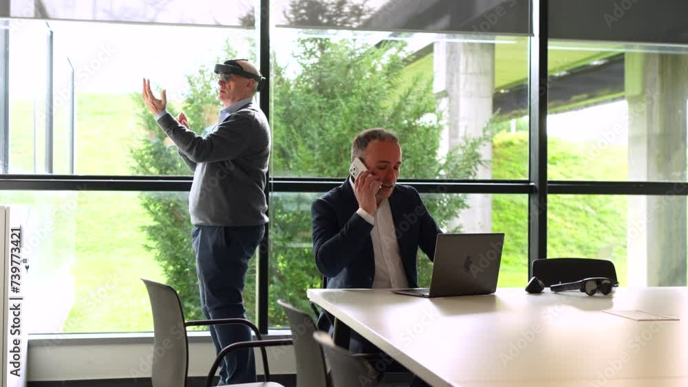 Businessman working using laptop while a colleague is using VR googles in the office Businessman working using laptop while a colleague is using VR googles in the office