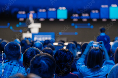 Back view of an attentive audience at a professional event with a speaker presenting on stage.