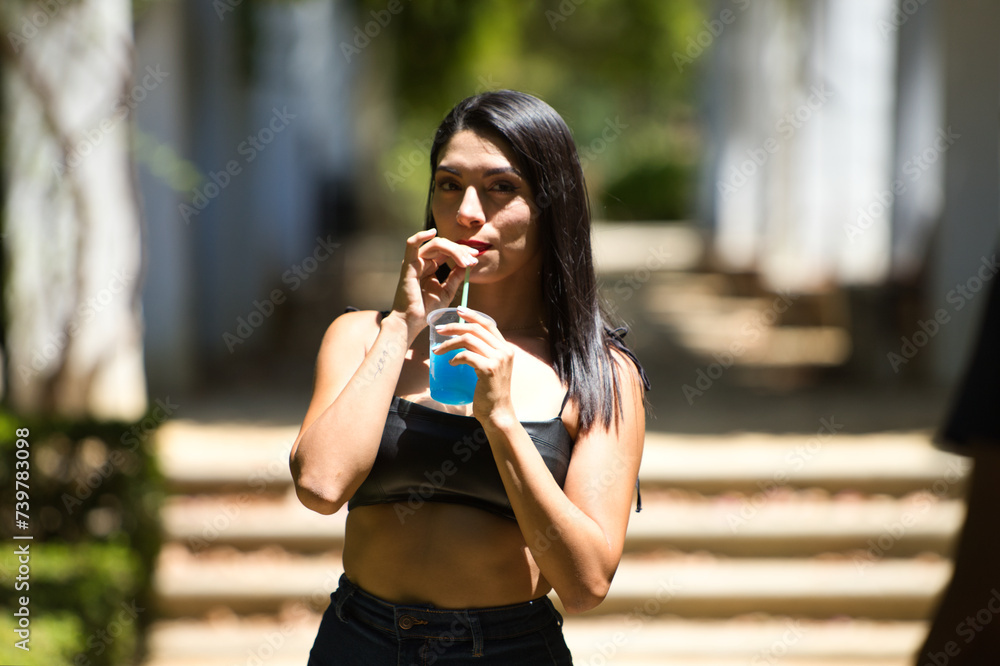 Young and beautiful Latin woman, cooling off from the heat, drinking a blue slushy drink through a straw. The woman drinks and shows her drink to the camera.