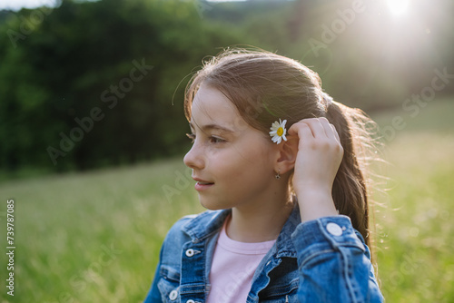 Portrait of beautiful young girl with flower behind ear  standing on meadow  enjoying warm spring day.