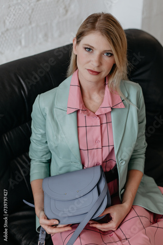 Elegant woman in a pink dress and mint jacket, holding a turquoise handbag and posing while sitting on the sofa