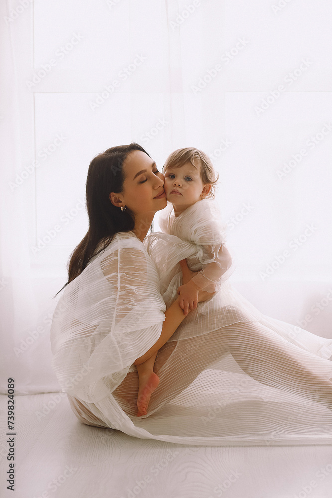 Studio portrait of happy mother sitting near window and hugging her baby girl. Mother and daughter in white dresses. Studio portrait of happy mother sitting near window and hugging her baby girl. Mother and daughter in white dresses.
