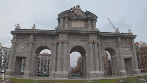 Dolly tracking shot of Puerta de Alcala in Madrid, Spain. Cloudy winter day with foregorund of grass and flowers wide angle panoramic view