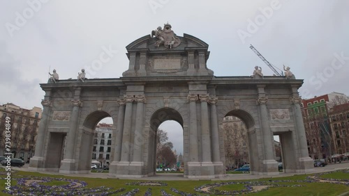 Dolly tracking shot of Puerta de Alcala in Madrid, Spain. Cloudy winter day with foregorund of grass and flowers wide angle panoramic view