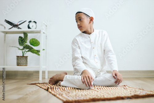 Muslim kid wearing skullcap praying salat in sitting position, reciting the salam facing the right direction photo