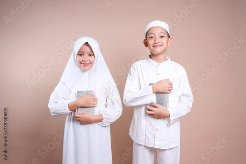 Portrait of Asian little boy and girl wearing hijab, holding holy book of Quran on their chest 