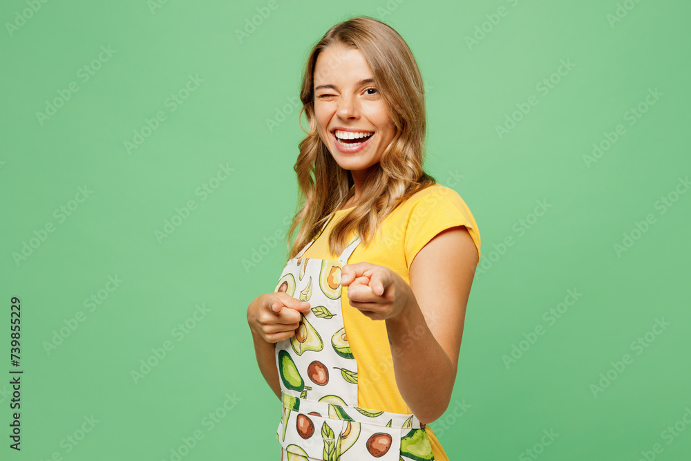 Young cheerful housewife housekeeper chef cook baker woman wear apron yellow t-shirt point index finger camera on you blink eye isolated on plain pastel green background studio. Cooking food concept. Young cheerful housewife housekeeper chef cook baker woman wear apron yellow t-shirt point index finger camera on you blink eye isolated on plain pastel green background studio. Cooking food concept.
