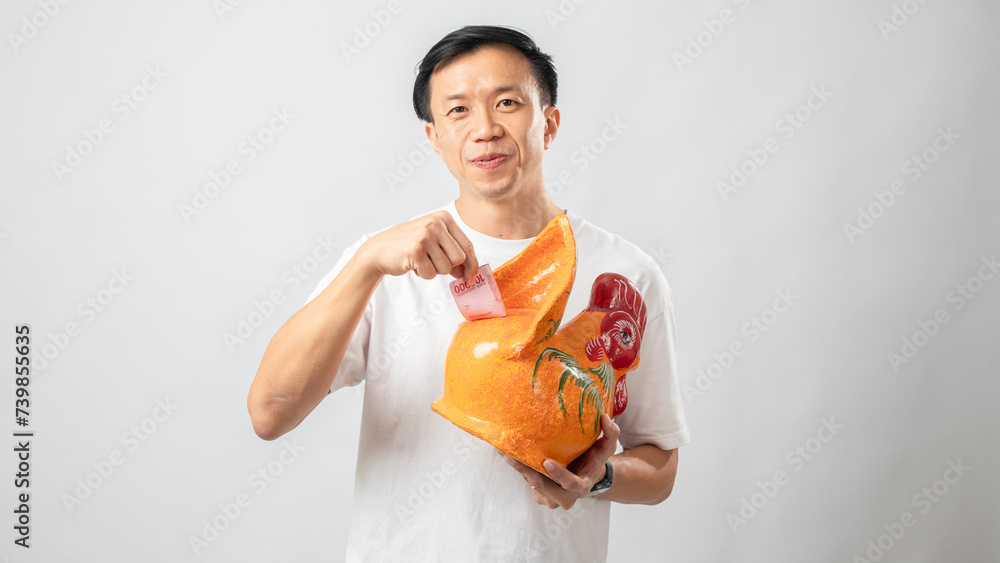 Portrait of an Indonesian Asian man, wearing a white T-shirt, posing while saving money with a chicken-shaped piggy bank, isolated on a white background. Portrait of an Indonesian Asian man, wearing a white T-shirt, posing while saving money with a chicken-shaped piggy bank, isolated on a white background.