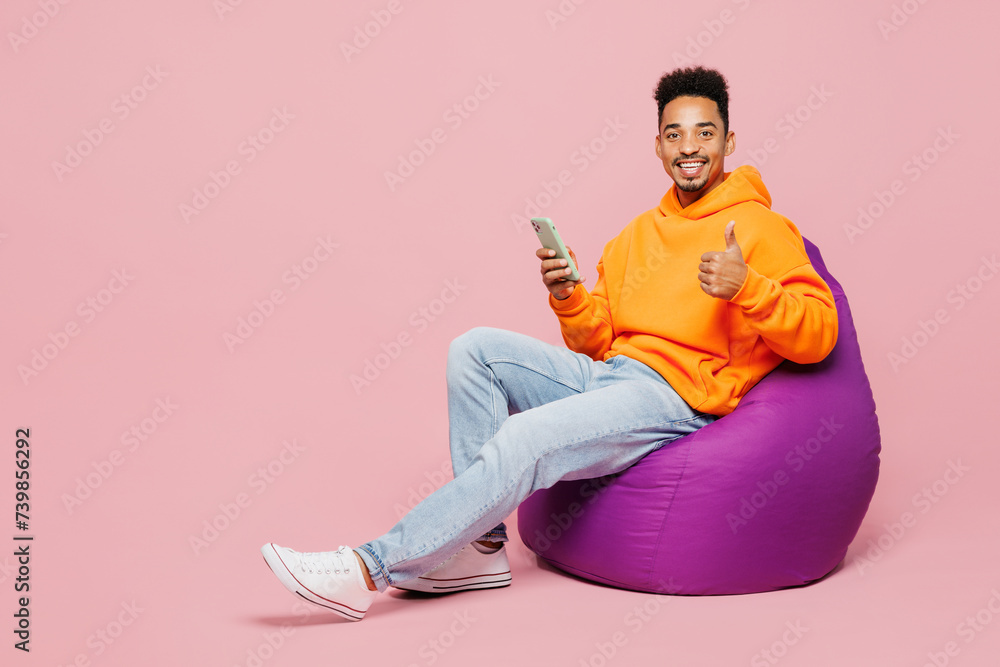 Full body young man of African American ethnicity wear yellow hoody casual clothes sit in bag chair hold in hand use mobile cell phone isolated on plain pastel light pink background Lifestyle concept Full body young man of African American ethnicity wear yellow hoody casual clothes sit in bag chair hold in hand use mobile cell phone isolated on plain pastel light pink background Lifestyle concept