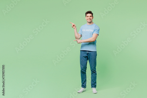 Full body smiling young man wears blue t-shirt title volunteer point index finger aside on area mock up isolated on plain green background. Voluntary free work assistance help charity grace concept.