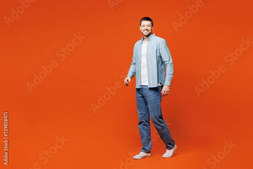 Full body side profile view young smiling fun man he wears blue shirt white t-shirt casual clothes walking going look camera isolated on plain red orange background studio portrait. Lifestyle concept.