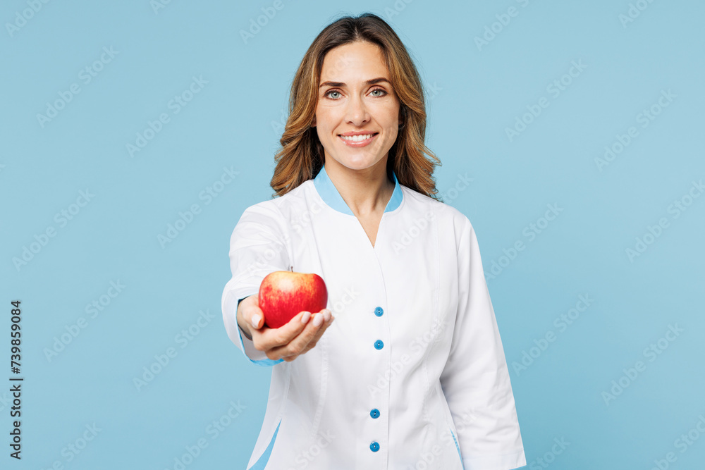 Female smiling happy doctor woman wears white medical gown suit work in hospital clinic office hold in hand giving apple fruit isolated on plain blue background studio. Health care medicine concept. Female smiling happy doctor woman wears white medical gown suit work in hospital clinic office hold in hand giving apple fruit isolated on plain blue background studio. Health care medicine concept.