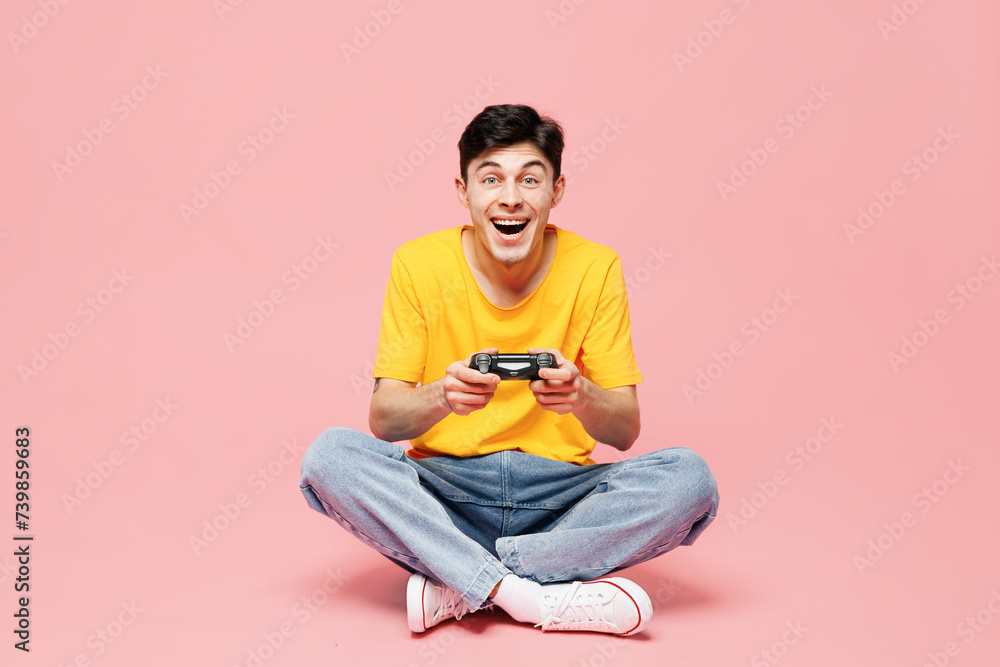 Full body happy young man he wears yellow t-shirt casual clothes sits hold in hand play pc game with joystick console isolated on plain pastel light pink background studio portrait. Lifestyle concept. Full body happy young man he wears yellow t-shirt casual clothes sits hold in hand play pc game with joystick console isolated on plain pastel light pink background studio portrait. Lifestyle concept.