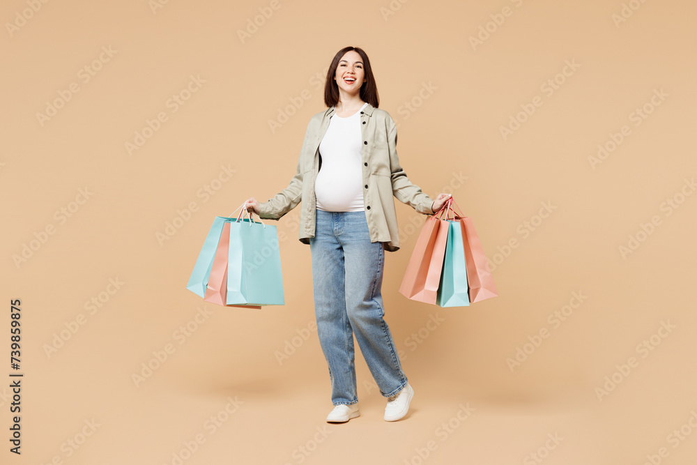 Full body smiling young pregnant woman future mom wear grey shirt with belly stomach tummy with baby hold shopping package bags isolated on plain beige background. Maternity family pregnancy concept. Full body smiling young pregnant woman future mom wear grey shirt with belly stomach tummy with baby hold shopping package bags isolated on plain beige background. Maternity family pregnancy concept.