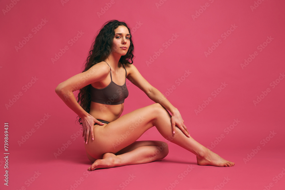 Curly, brunette woman sitting on floor and posing looking at camera wearing brown lingerie against pink studio background. Concept of wellness, female health, healthy lifestyle, cosmetology, spa.