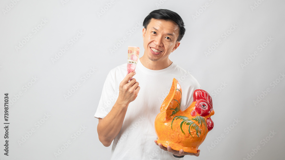 Portrait of an Indonesian Asian man, wearing a white T-shirt, posing while saving money with a chicken-shaped piggy bank, isolated on a white background. Portrait of an Indonesian Asian man, wearing a white T-shirt, posing while saving money with a chicken-shaped piggy bank, isolated on a white background.