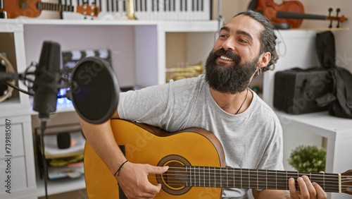 A cheerful hispanic man plays guitar in a home studio with microphones and instruments visible.