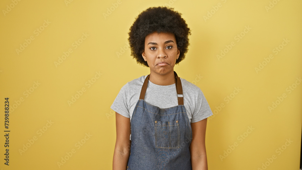 African american woman with curly hair in a denim apron standing against a yellow background African american woman with curly hair in a denim apron standing against a yellow background