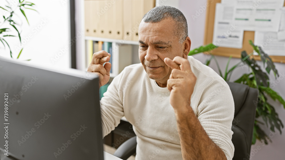 Fototapeta premium Middle-aged hispanic man crossing fingers for luck while working at his office computer indoors.