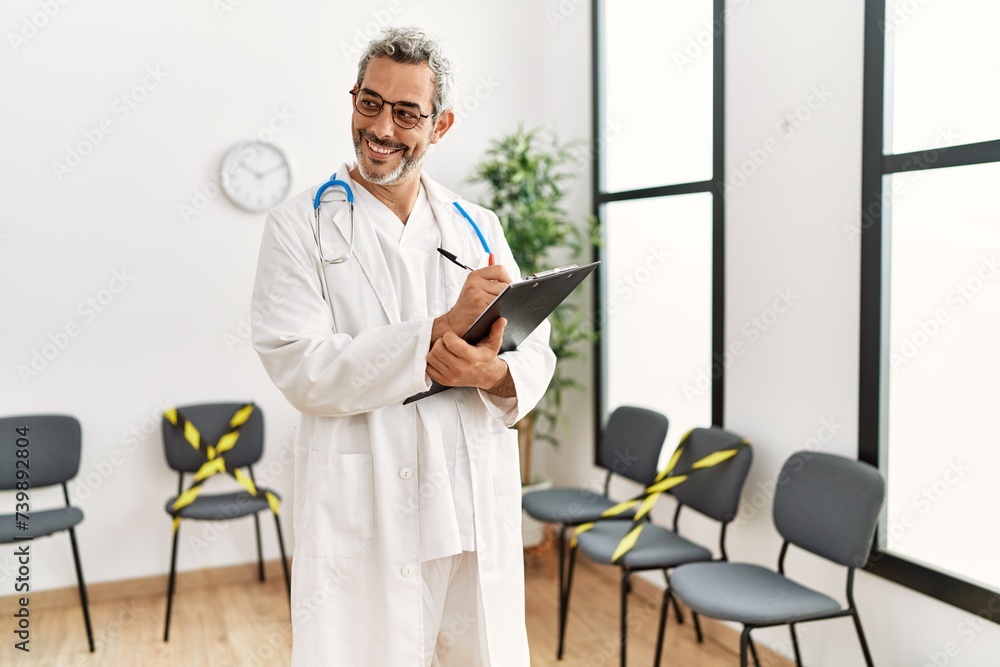Middle age grey-haired man doctor smiling confident writing medical report at clinic waiting room