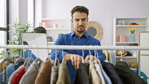 Εκτύπωση καμβά Handsome hispanic man with beard selecting clothes in a bright indoor room, reflecting fashion and wardrobe choices