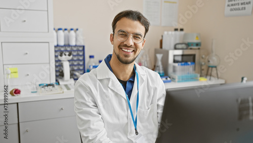 A smiling young hispanic man with a beard wearing glasses and a lab coat in a laboratory setting.