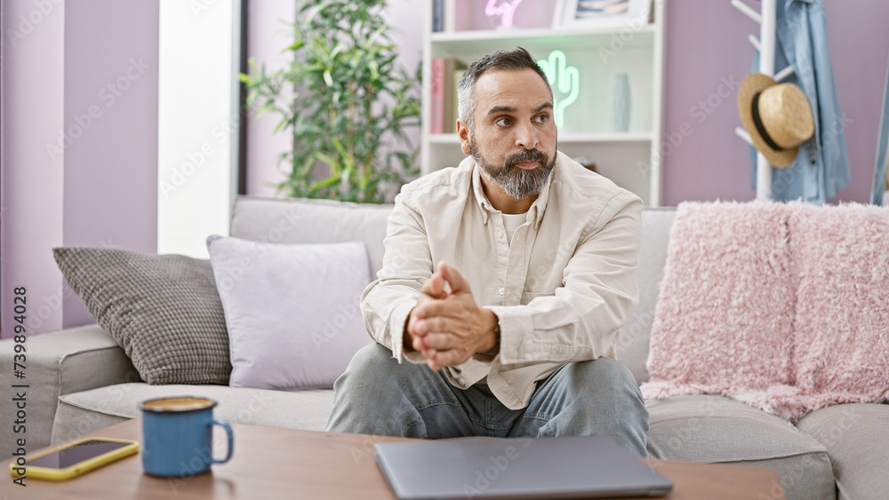 Grey-haired mature man with a beard in a casual indoor setting, sitting thoughtfully on a sofa with laptop and coffee. Grey-haired mature man with a beard in a casual indoor setting, sitting thoughtfully on a sofa with laptop and coffee.