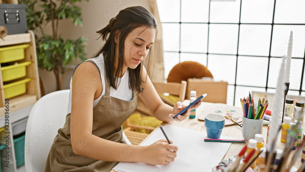 Enthralling glimpse of a beautiful young hispanic woman artist, earnestly drawing on her notebook, armed with a smartphone in hand at an art studio. Enthralling glimpse of a beautiful young hispanic woman artist, earnestly drawing on her notebook, armed with a smartphone in hand at an art studio.