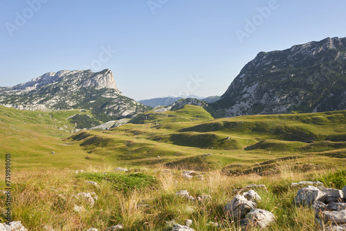 Serene green slopes ascend towards a clear sky, hinting at the majesty of the mountainous terrain. The lush grasses and stratified rock formations invite adventure and exploration
