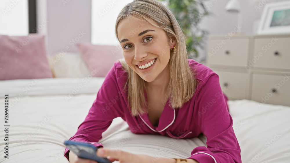 Smiling woman in pink pajamas holding smartphone in a bright bedroom Smiling woman in pink pajamas holding smartphone in a bright bedroom